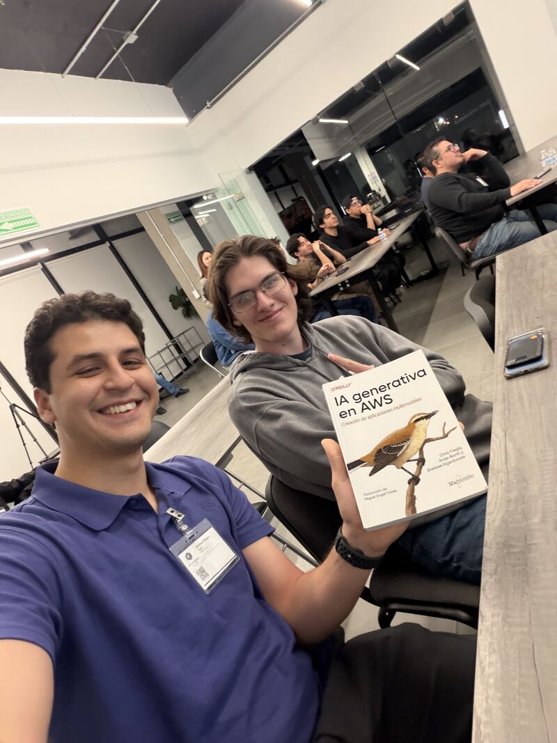 Dos miembros del AWS User Group Hermosillo sonriendo en un encuentro, sosteniendo un ejemplar del libro IA generativa en AWS (O'Reilly), con más asistentes sentados al fondo