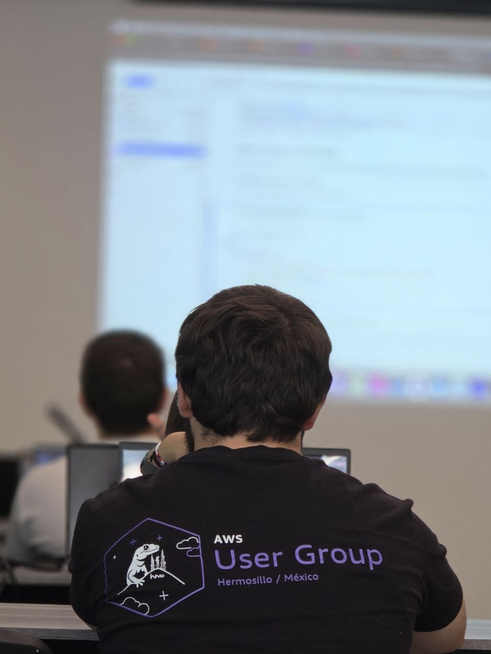 Persona de espaldas en un taller del AWS User Group Hermosillo, con la camiseta oficial de la comunidad frente a una pantalla de trabajo