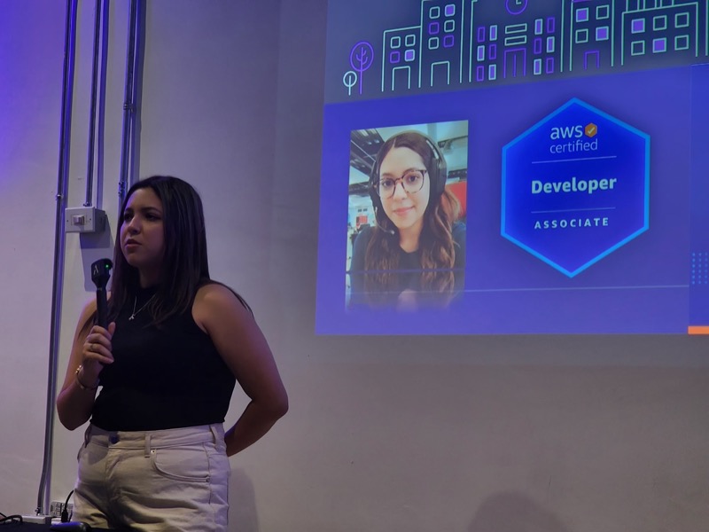 Presentadora en el escenario junto a la proyección de una speaker invitada por videollamada y su insignia AWS Certified Developer Associate