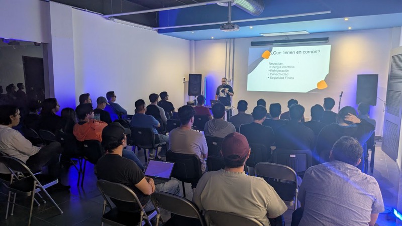 Charla sobre infraestructura de cómputo ante una sala llena, diapositiva "¿Qué tienen en común? Necesitan: energía eléctrica, refrigeración, conectividad, seguridad física"